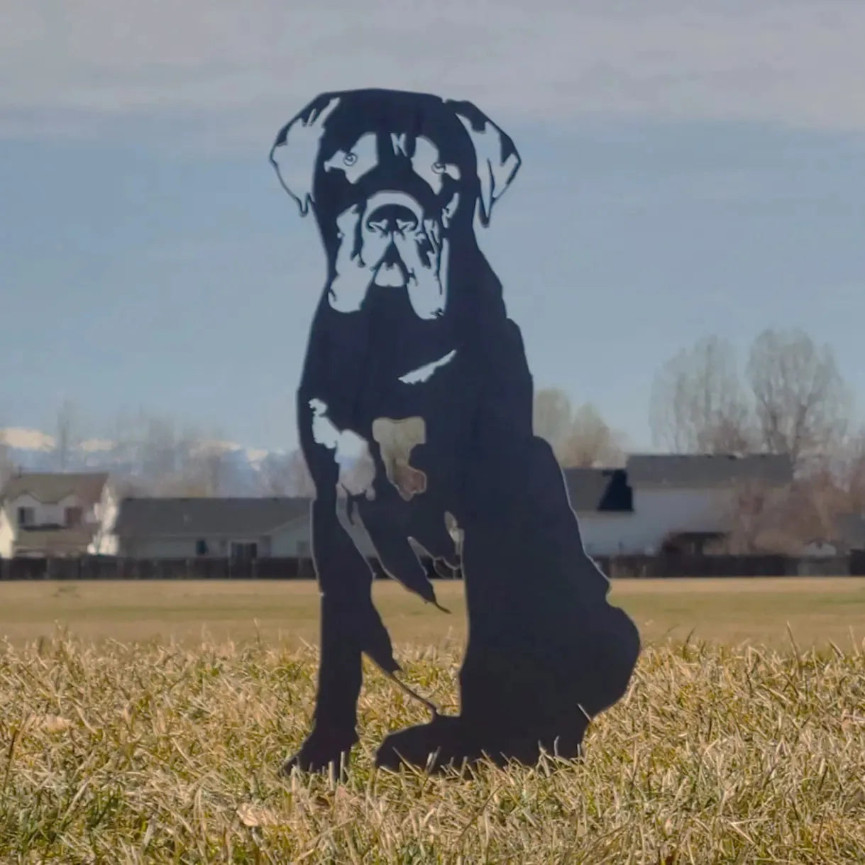 Mastiff metal silhouette in black, sitting on grass with a clear blue sky and distant homes in the background.