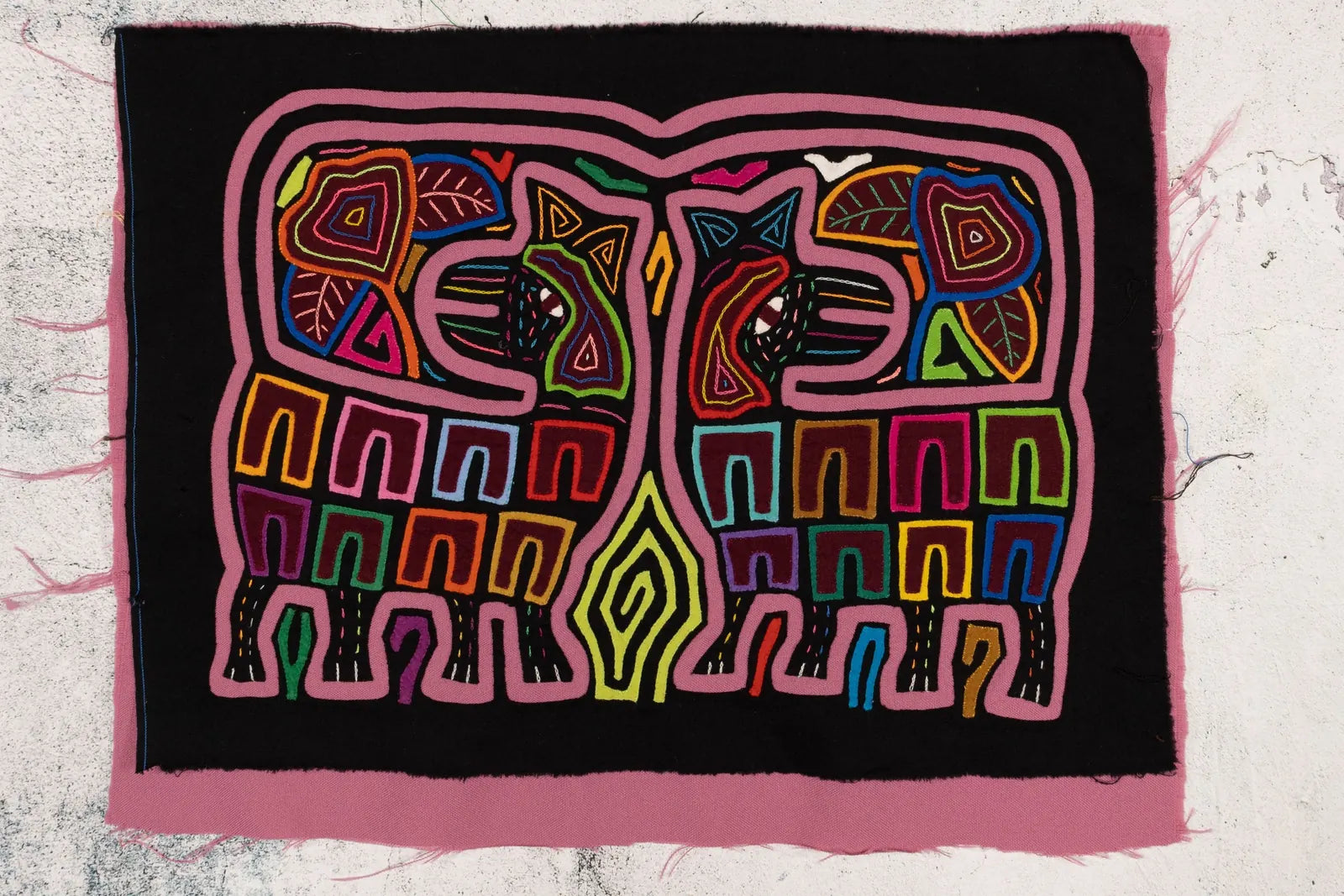 Hand-stitched Mola tapestry featuring vibrant geometric patterns and animal designs on black fabric with a pink border.