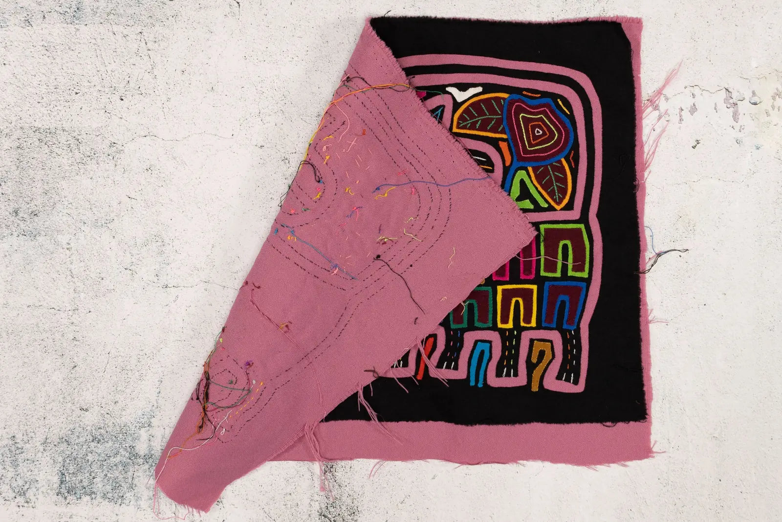 Hand-stitched Mola textile featuring vibrant geometric patterns on a black background, with soft pink fabric exposed.
