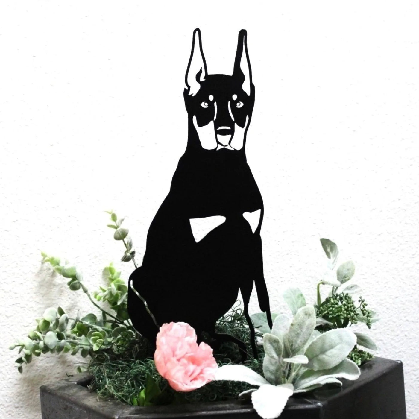 Black metal silhouette of a Miniature Pinscher surrounded by greenery and flowers in a planter.