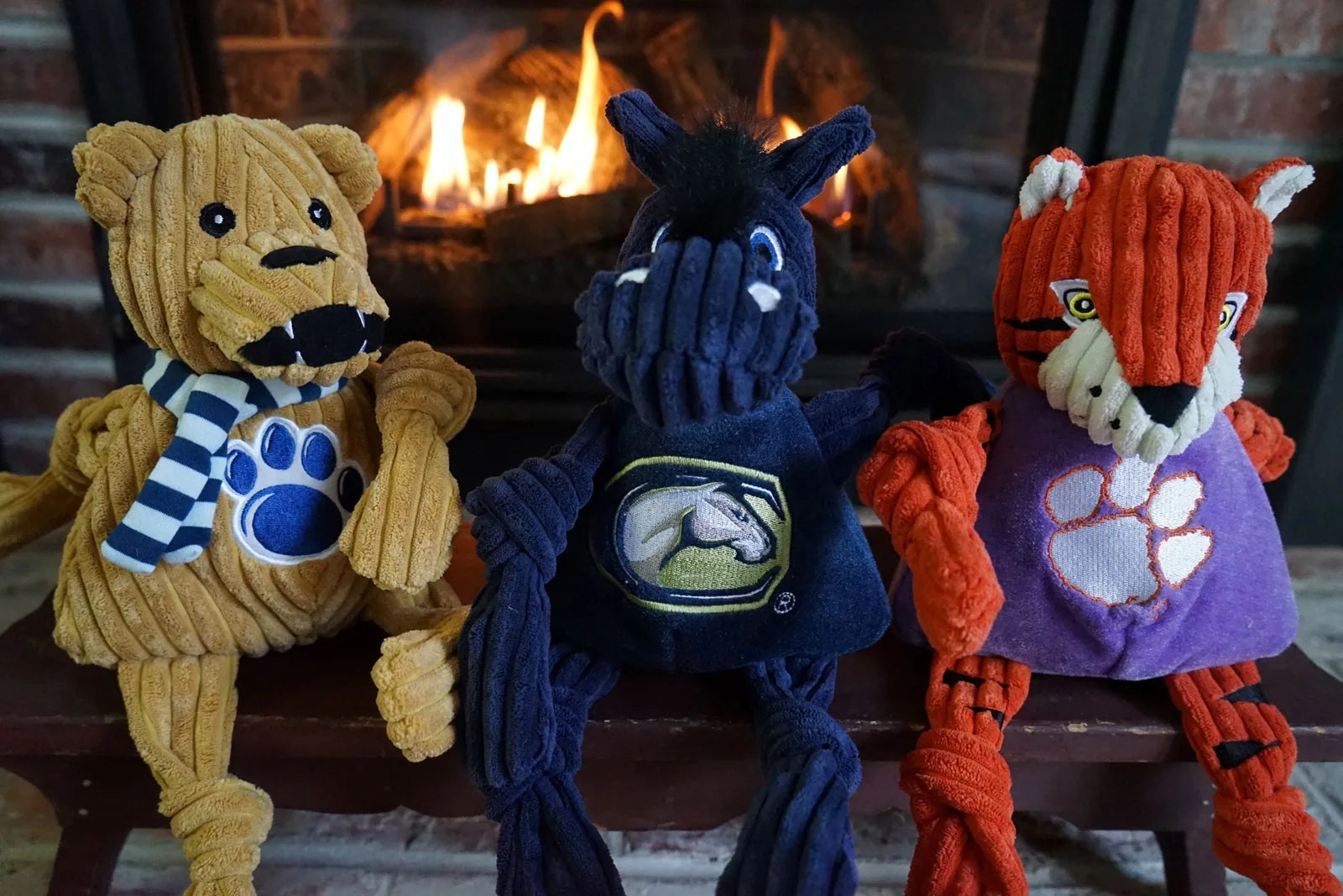 Plush Penn State Nittany Lion toy in gold corduroy with knotted limbs and striped scarf, displayed in front of a fireplace.