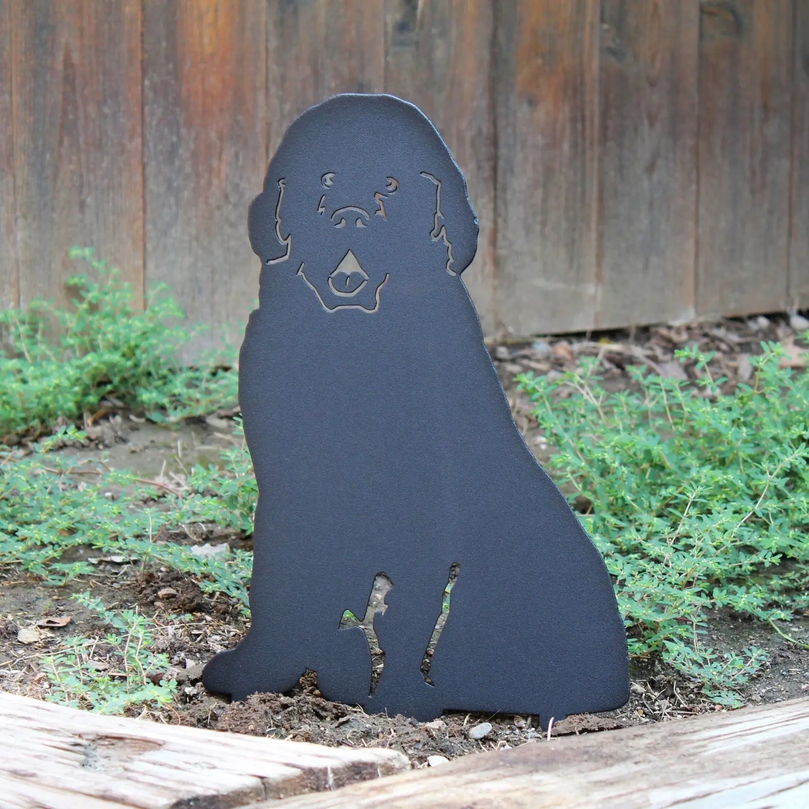 Solid black metal silhouette of a Newfoundland dog, partially embedded in garden soil, surrounded by green foliage.
