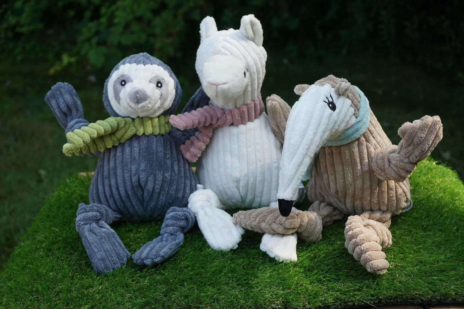 Plush dog toys featuring a blue sloth, white llama, and brown anteater, each with ribbed texture and colorful scarves.