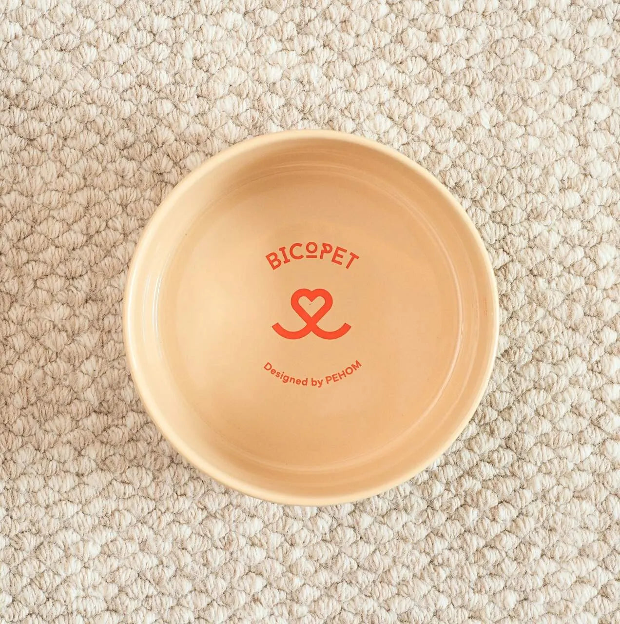 Ceramic Nomnom Bowl in soft beige with red "BICOPET" design, resting on a textured neutral surface.