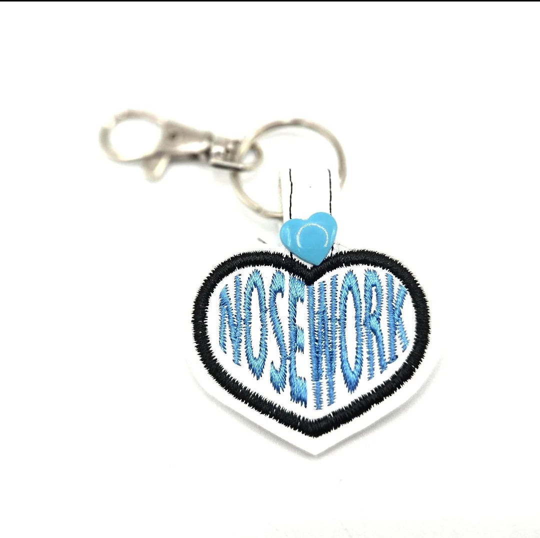 Embroidered heart keychain featuring "NOSEWORK" in blue with black outline and a light blue attachment clip.