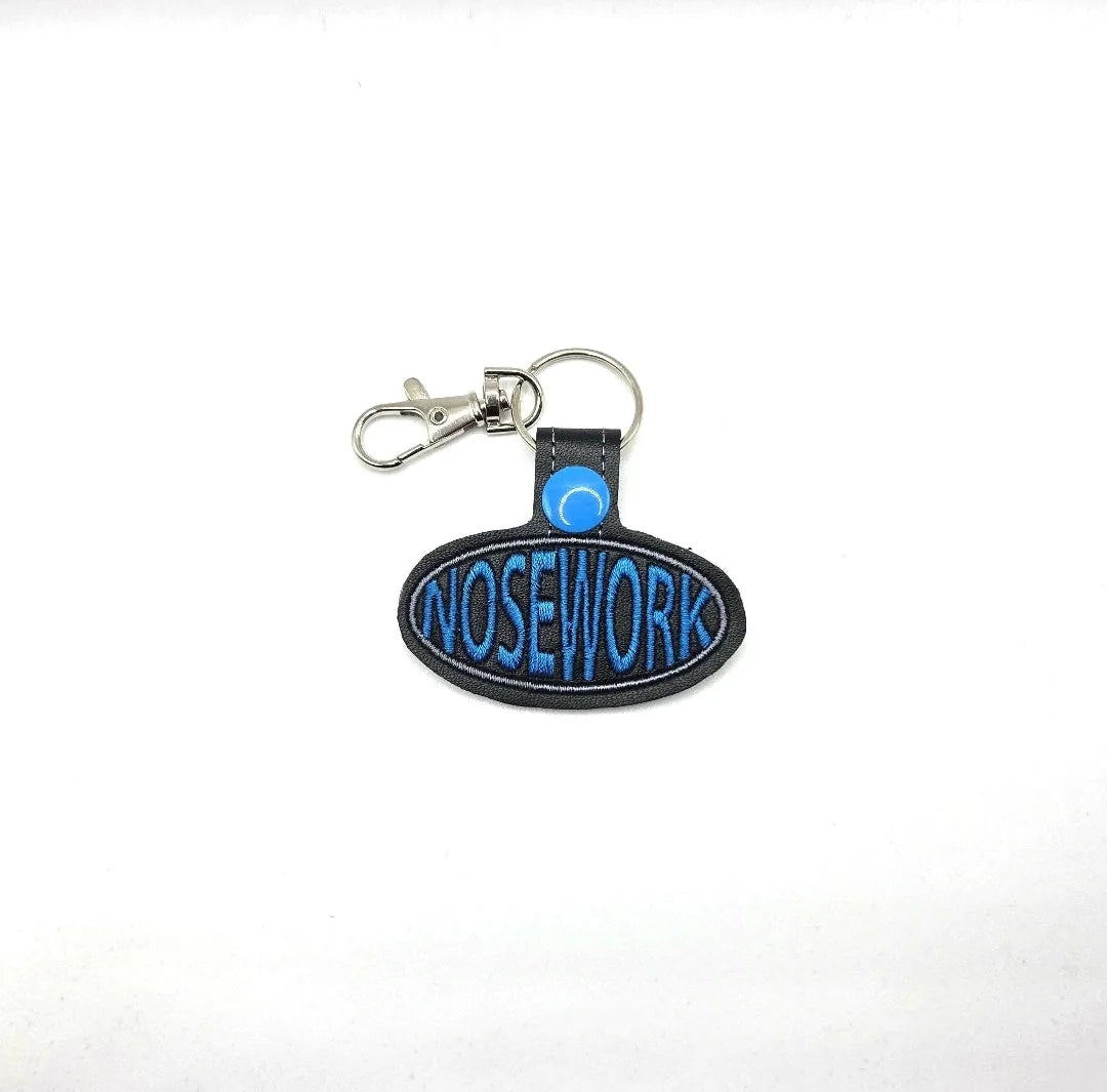 Embroidered oval keychain featuring "NOSEWORK" in blue on a black background with a silver swivel clip.