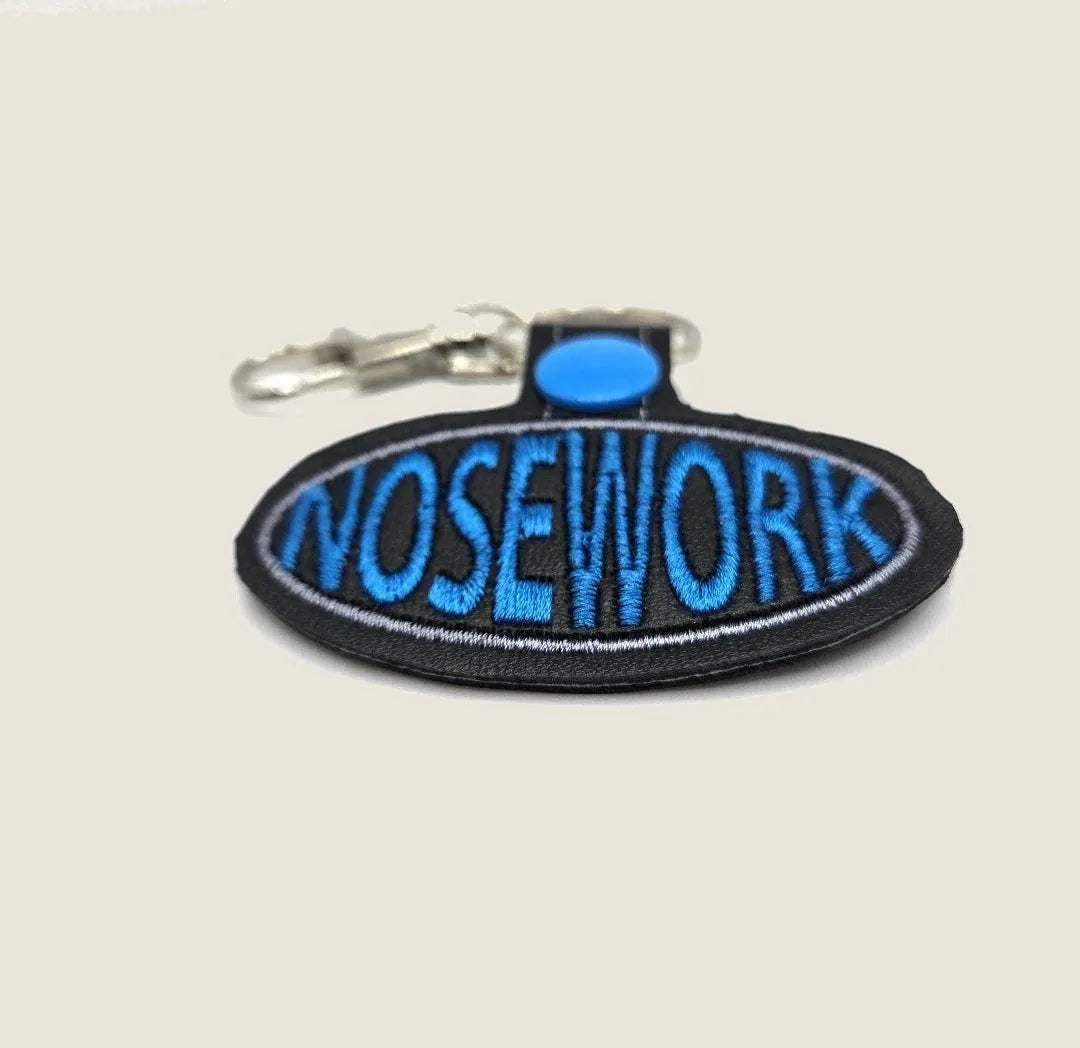 Embroidered keychain featuring "NOSEWORK" in bold blue on an oval black background, ideal for dog training enthusiasts.