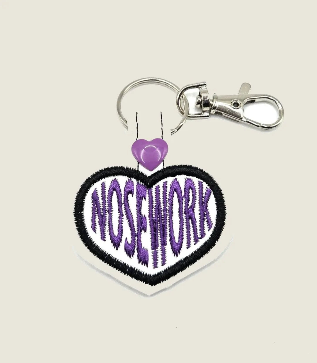Embroidered heart-shaped keychain with "NOSEWORK" in bold purple on a white background, attached with a silver clip.