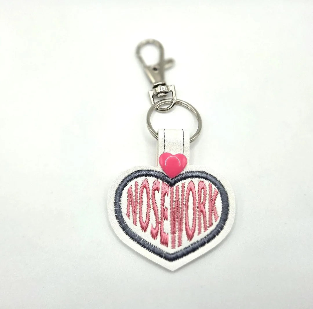 Heart-shaped keychain features "NOSEWORK" in pink embroidery with a pink heart, attached to a silver clip.