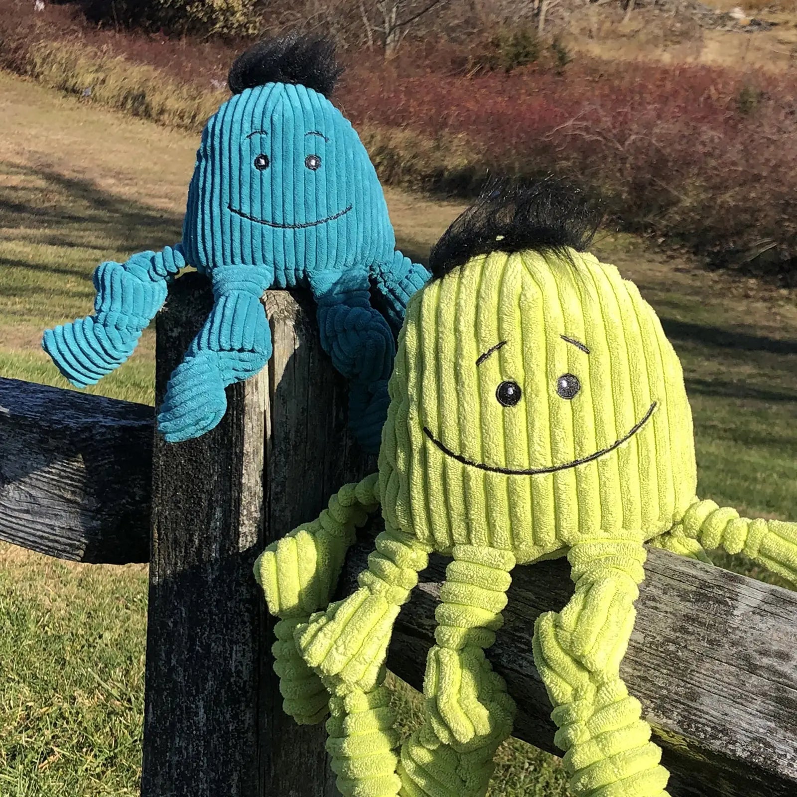 Colorful plush Octavie Octopus Knottie® toys in sea blue and octo green, smiling, with knotted limbs, on a wooden fence.