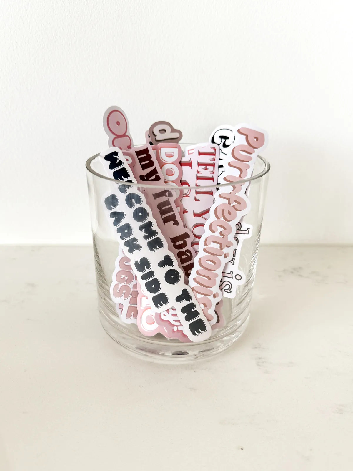 Colorful dog-themed stickers, featuring playful text, displayed in a clear glass container against a neutral background.