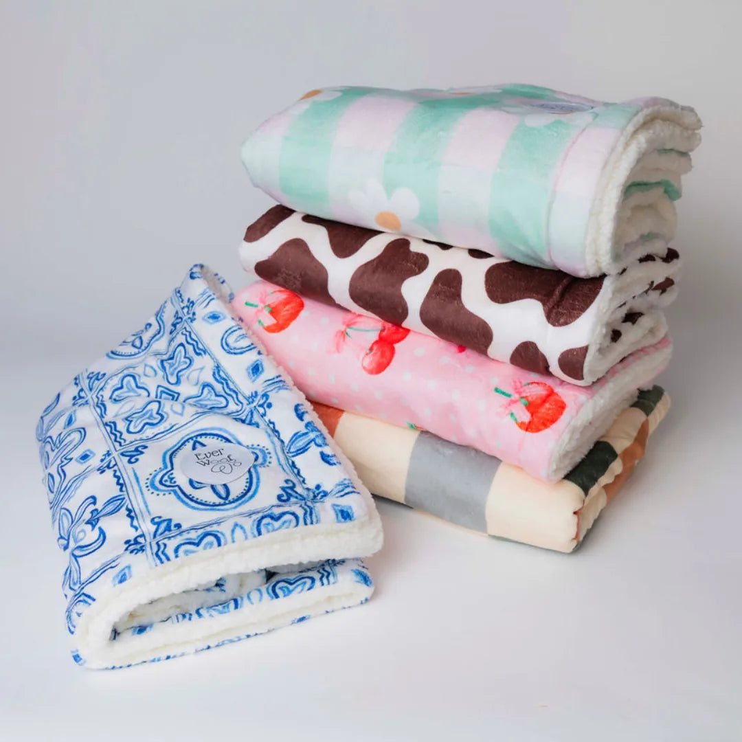 Colorful assorted dog blankets stacked, featuring floral, cow, and fruit patterns with soft micro-fiber and cream sherpa s...