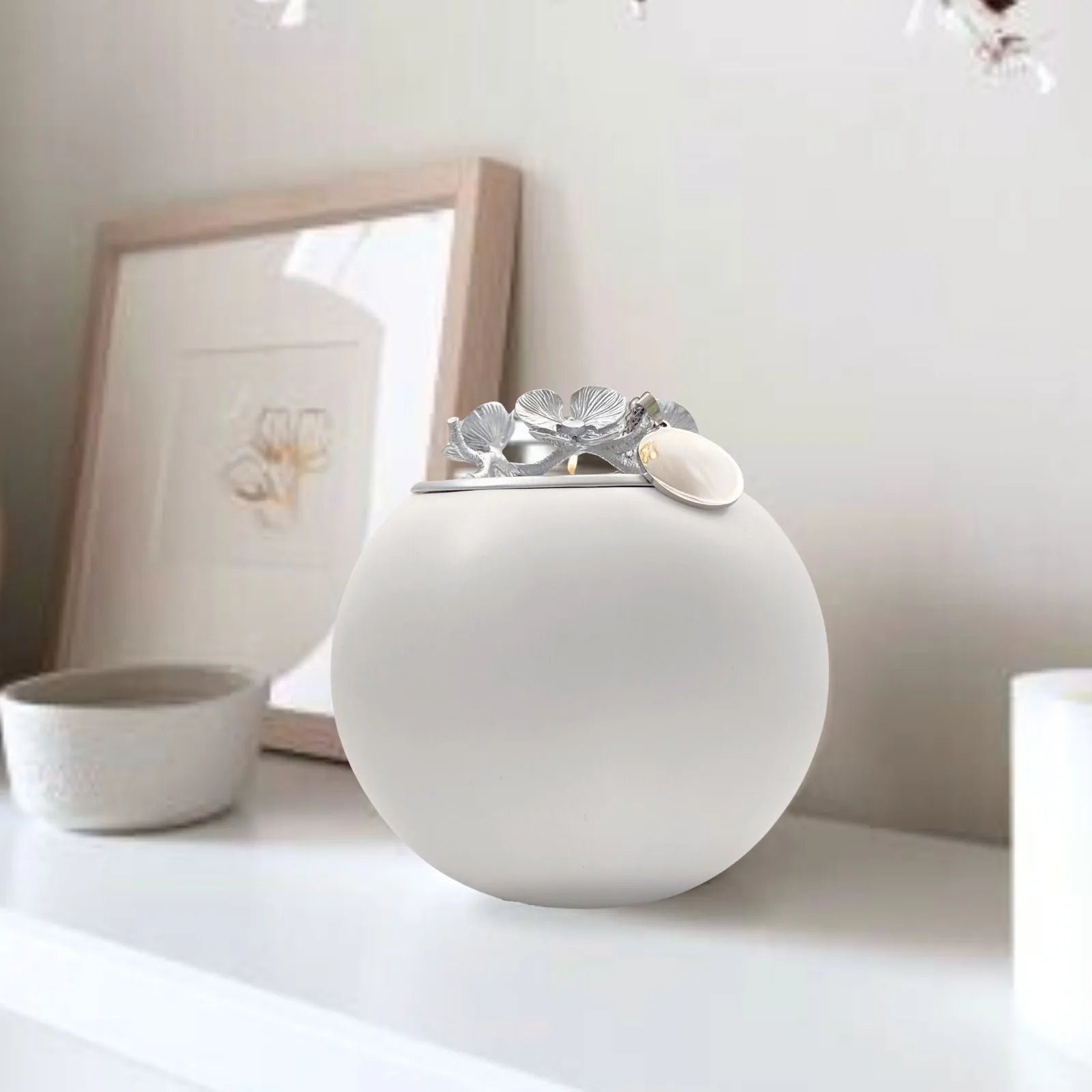 Handmade white orchid urn with silver floral lid, elegantly displayed on a shelf beside framed art and a small bowl.