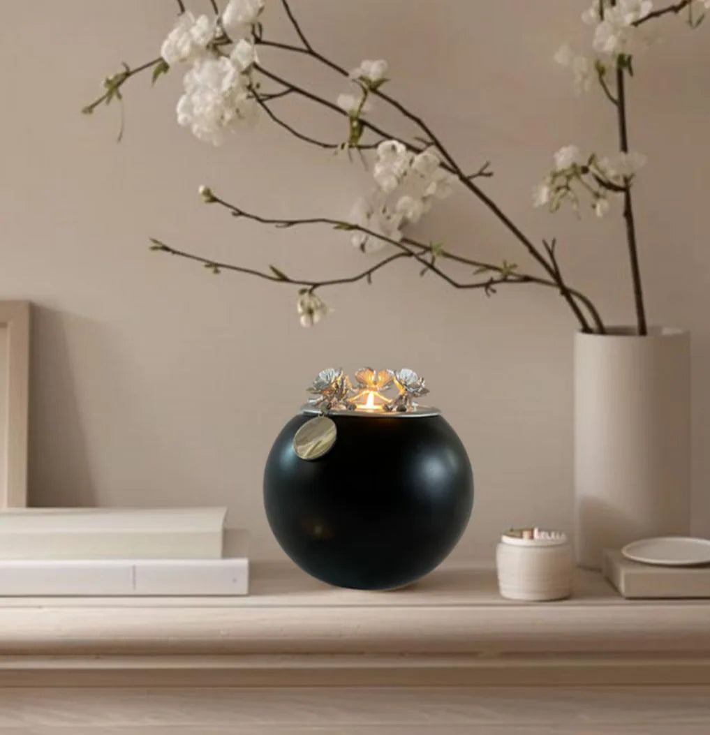 Handmade black orchid urn with floral silver lid, elegantly displayed on a soft wood shelf beside decorative items.
