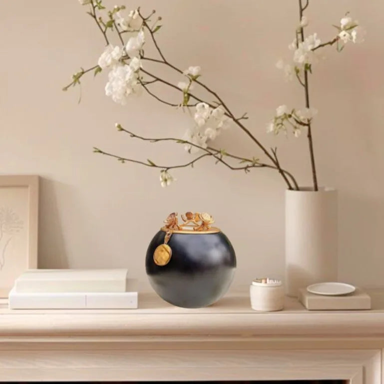Handmade black orchid urn with a gold lid and charm, elegantly displayed among decorative items on a wooden surface.
