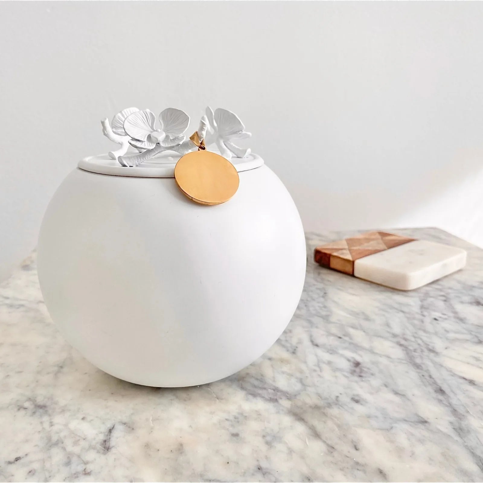 Handmade white keepsake urn with floral lid and gold charm, resting on a marble surface, ideal for memorializing loved ones.
