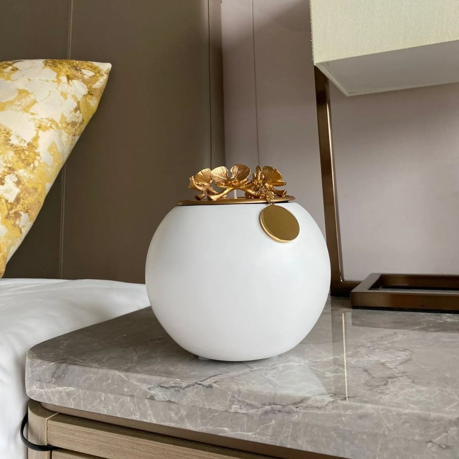 Handmade white urn with a gold floral lid, displayed on a marble top beside a decorative pillow.
