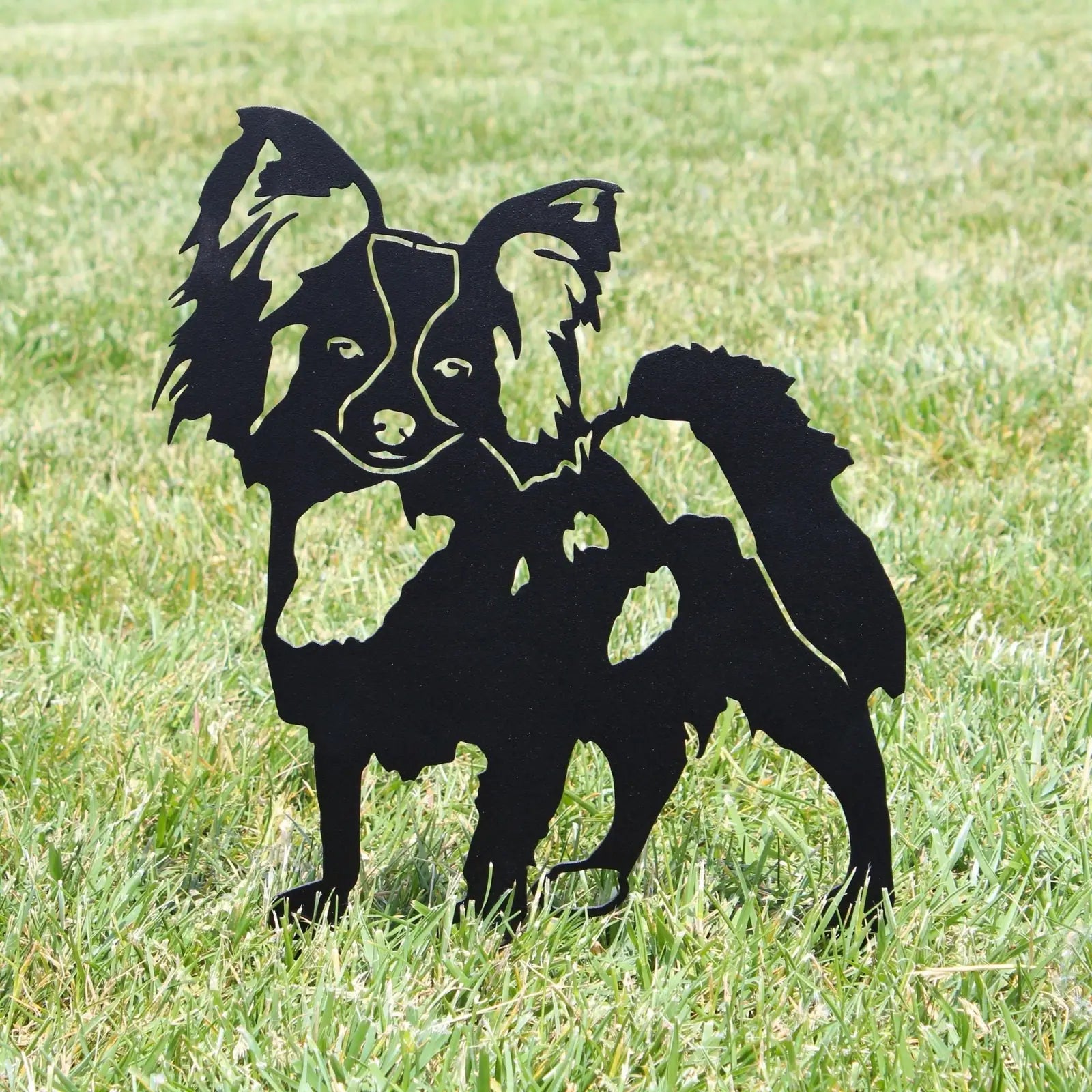 Black metal silhouette of a Papillon dog standing on green grass, showcasing detailed features and sturdy design.