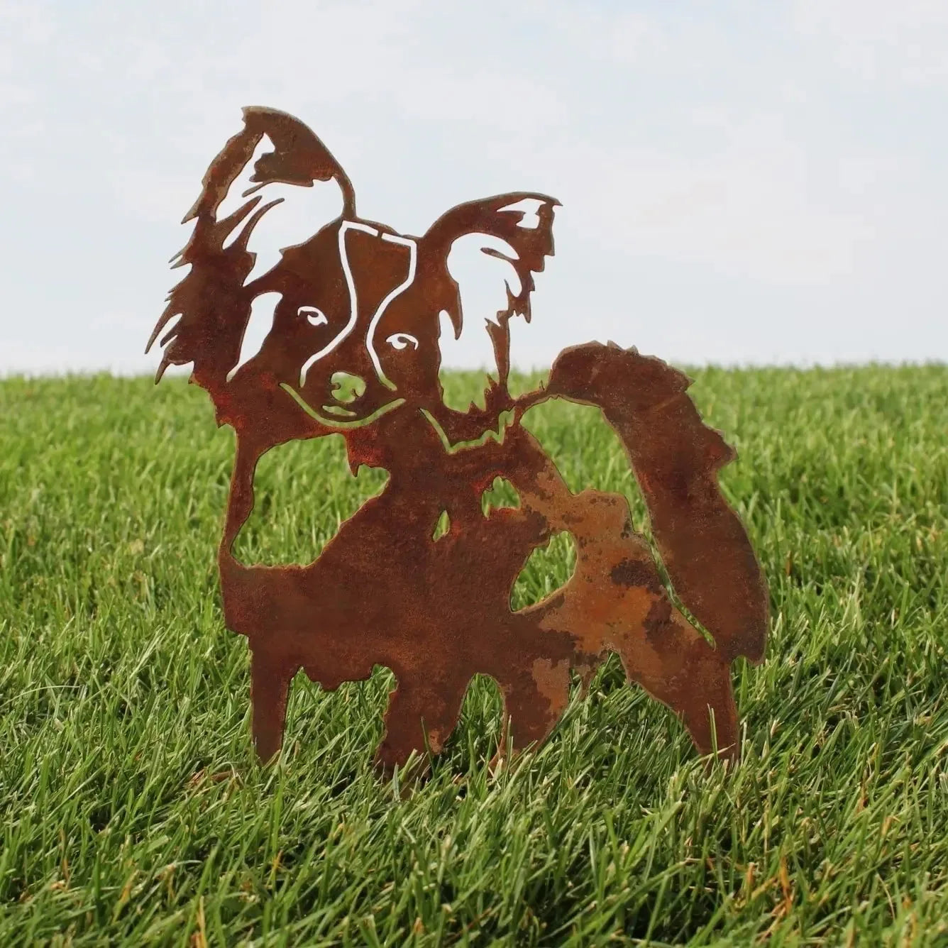 Rusty Corten steel silhouette of a Papillon dog, standing on green grass, showcasing intricate metal art design.