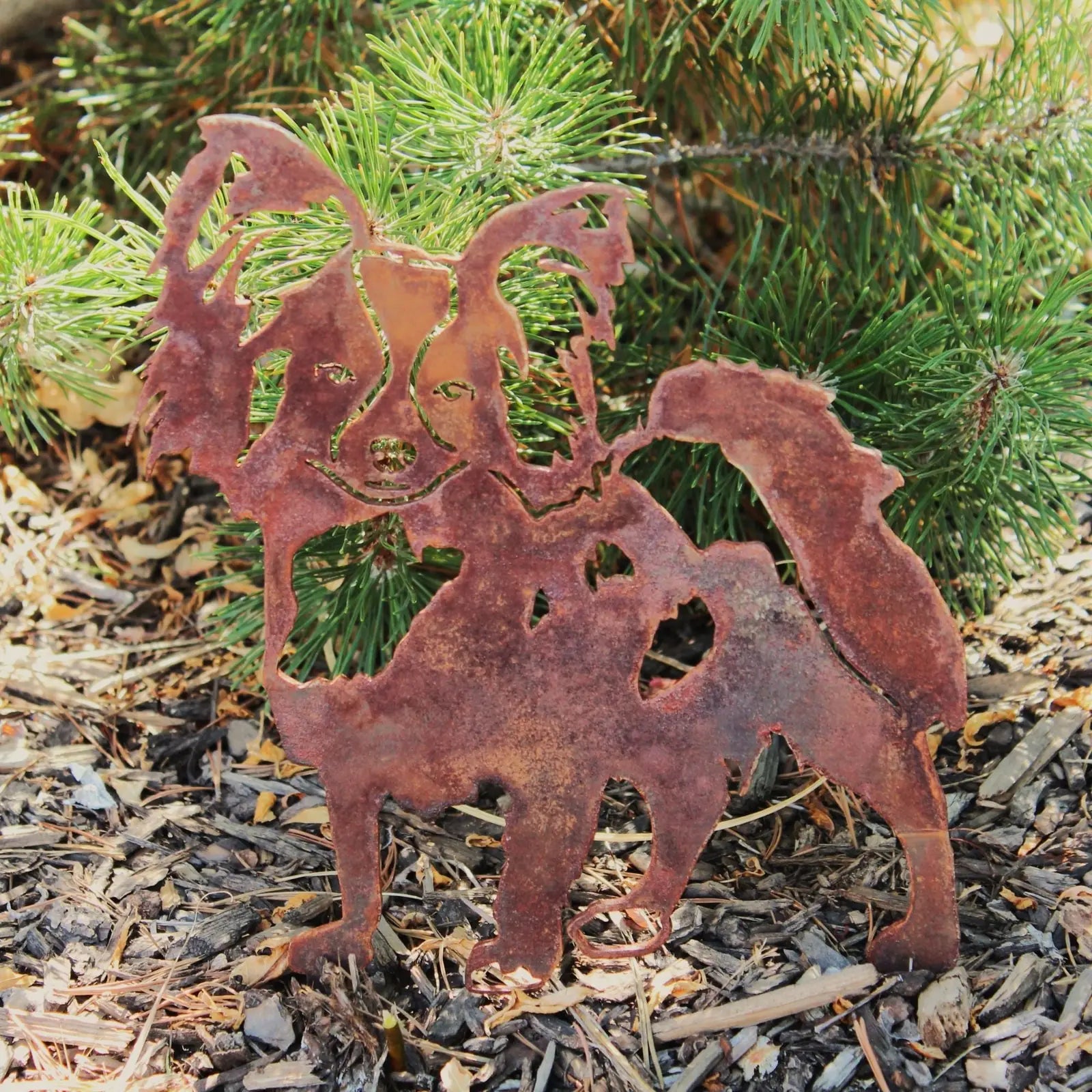 Rusty Corten steel Papillon dog silhouette garden decor, set against green pine needles and mulch. Ideal for outdoor spaces.