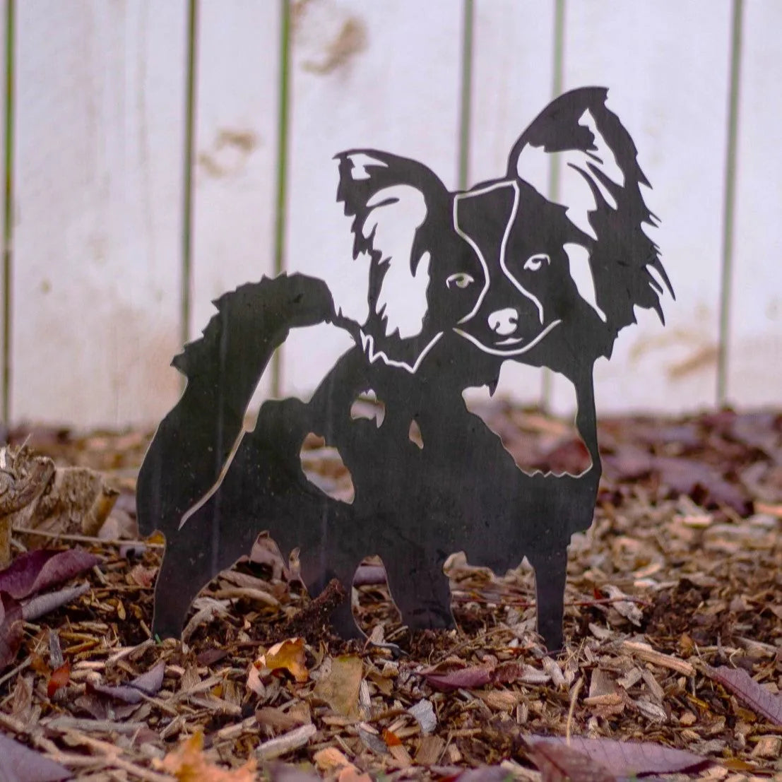 Black metal silhouette of a Papillon dog, set among brown mulch and leaves, perfect for garden decor.