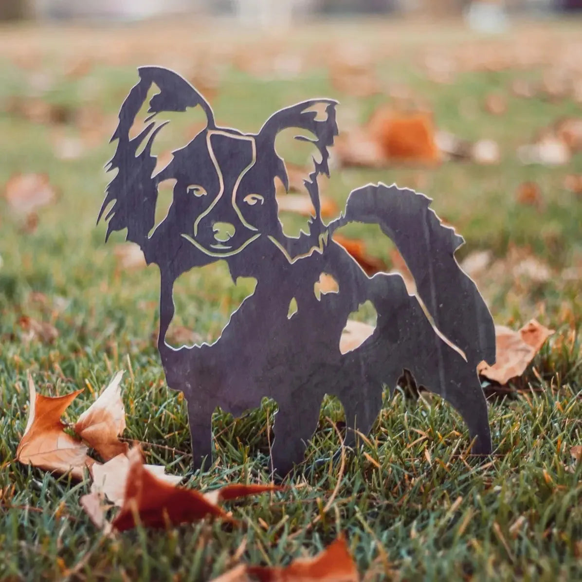 Handcrafted Papillon dog silhouette in matte black metal, set against grassy ground with autumn leaves.