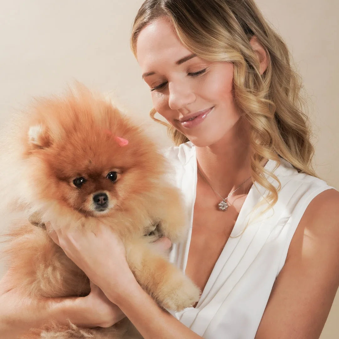 Delicate silver paw print cremation necklace worn by a woman holding a fluffy orange Pomeranian against a soft background.