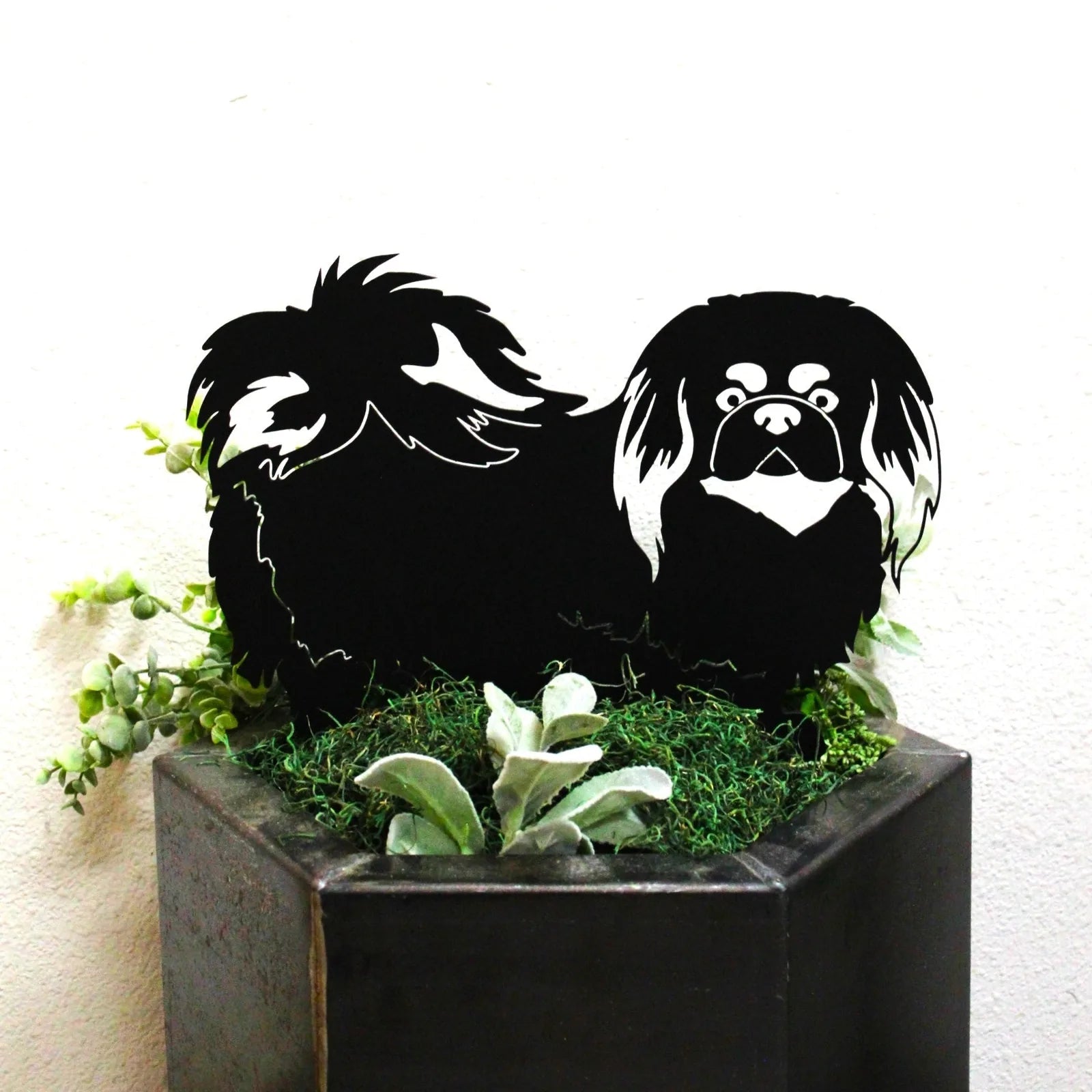 Black Pekingese metal silhouette art piece displayed in a planter with greenery, showcasing intricate detailing and design.