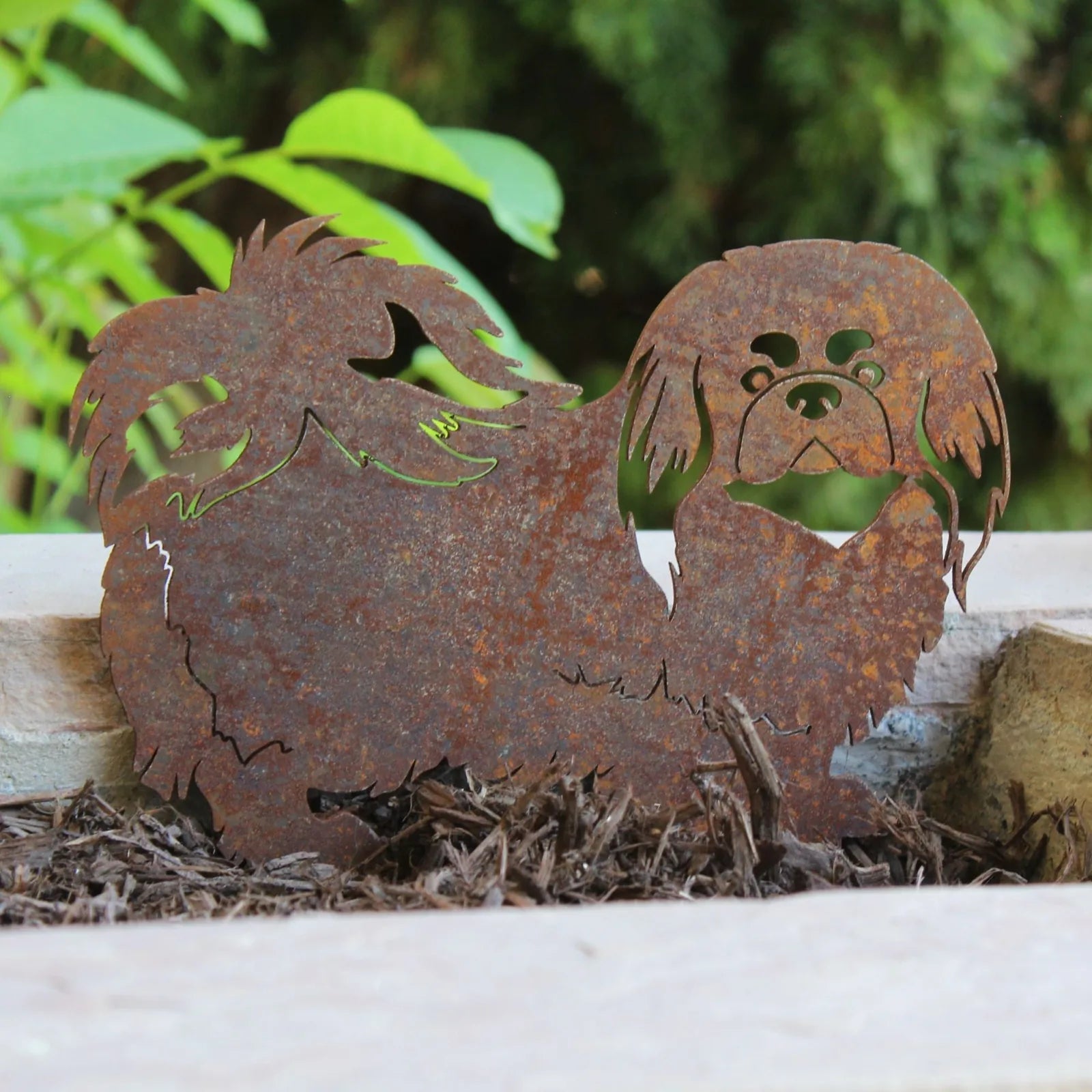 Handmade rust-colored Pekingese metal silhouette displayed among mulch, showcasing intricate detailing and weathered texture.