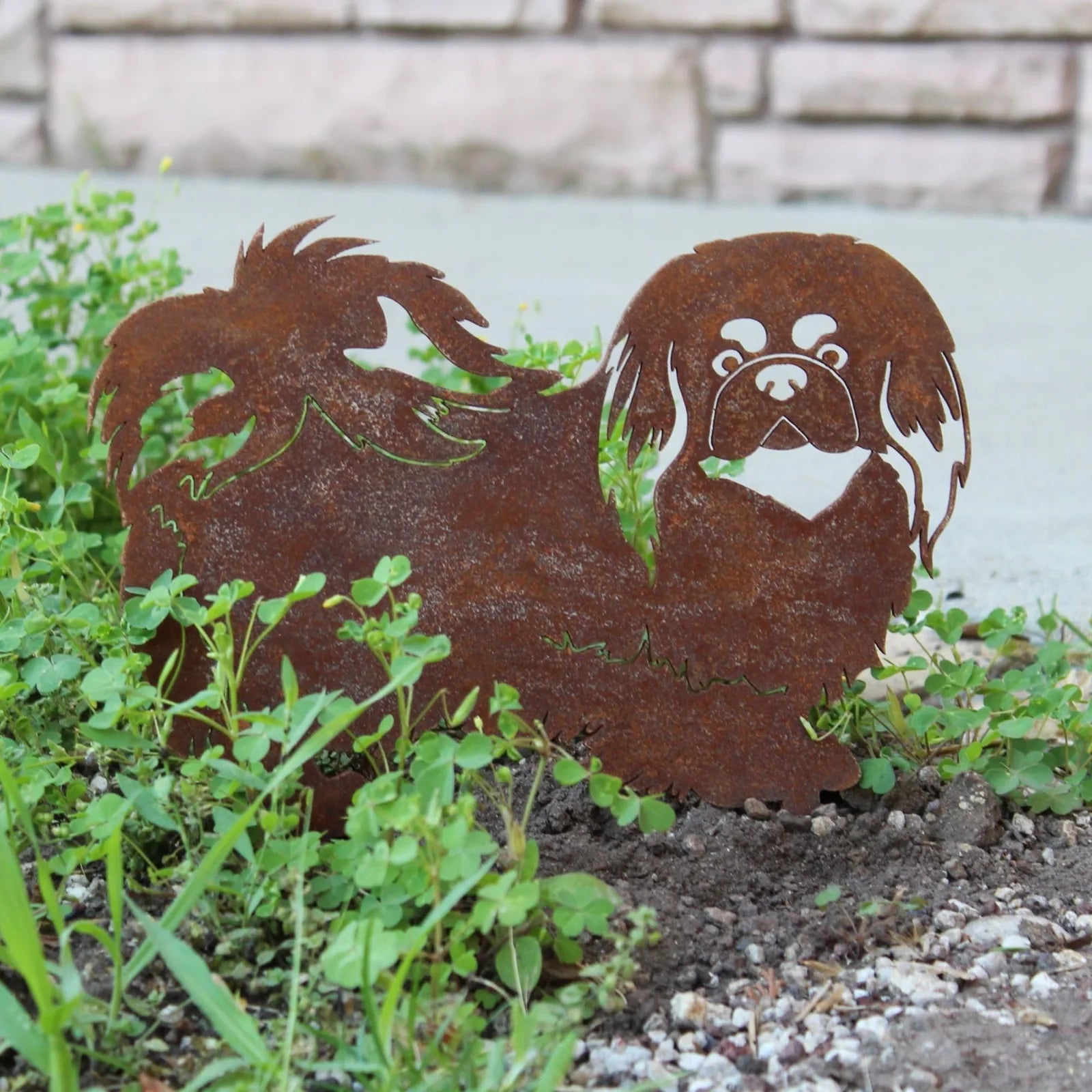 Handmade Pekingese metal silhouette in weathered rust finish, set among green ground cover plants.