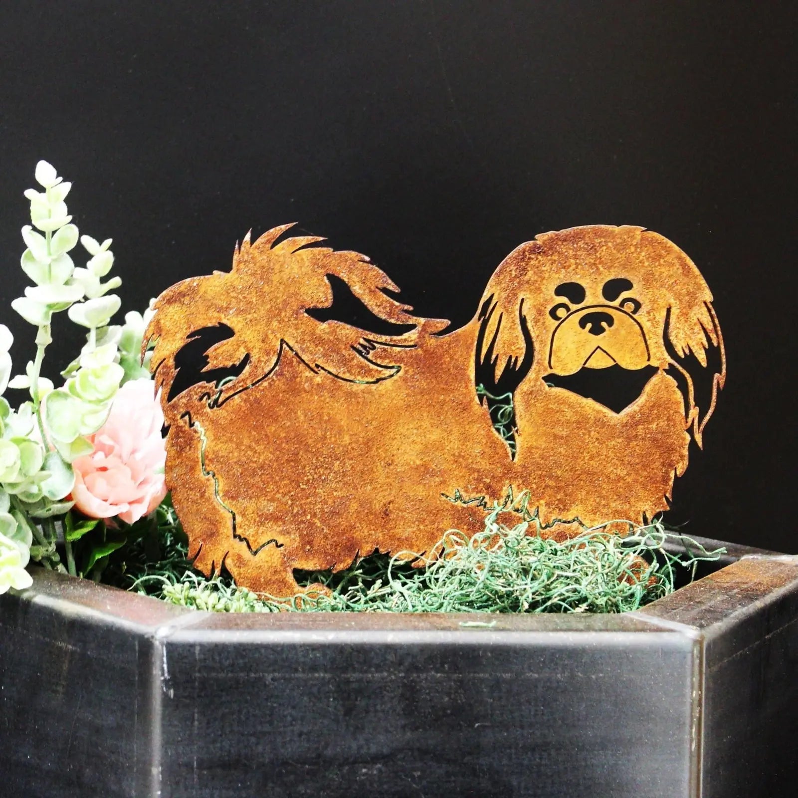 Rusty Corten steel silhouette of a Pekingese dog, surrounded by greenery and displayed in a black container.