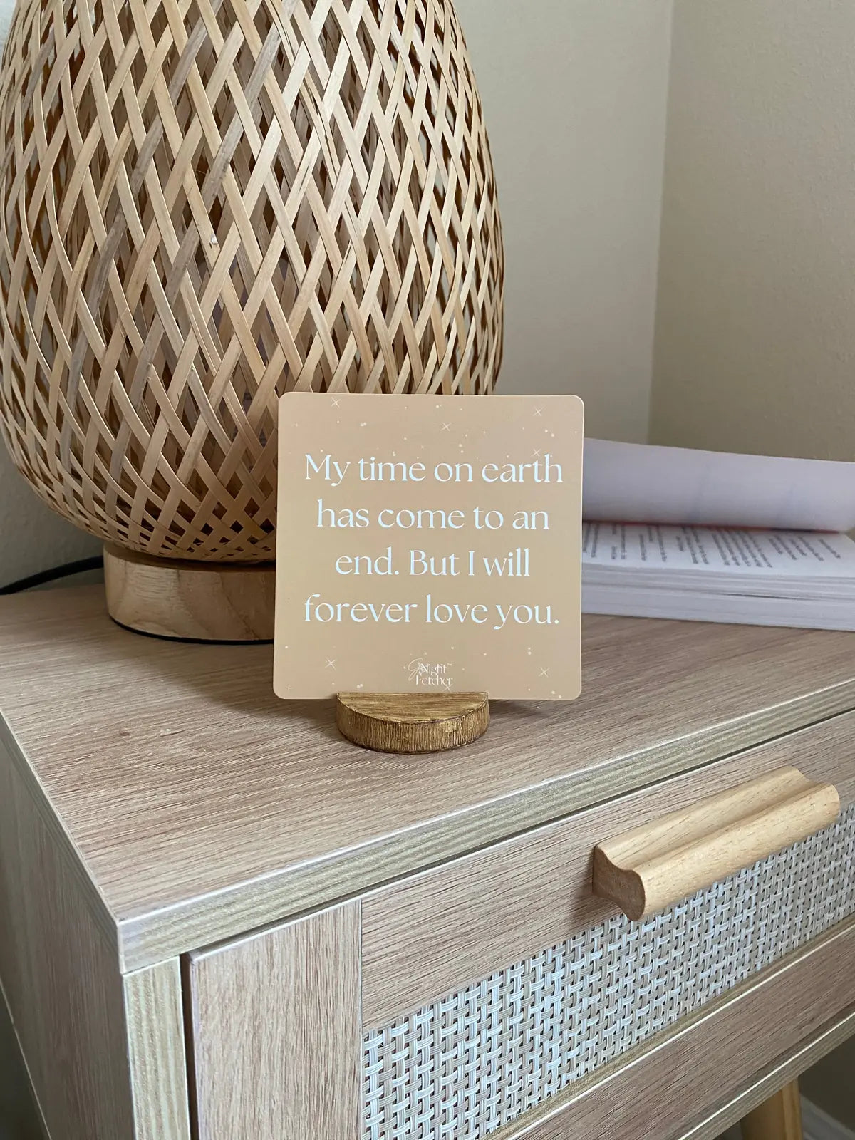 Sympathy card with a heartfelt message displayed on a wooden stand, beside a woven lamp on a light wood nightstand.