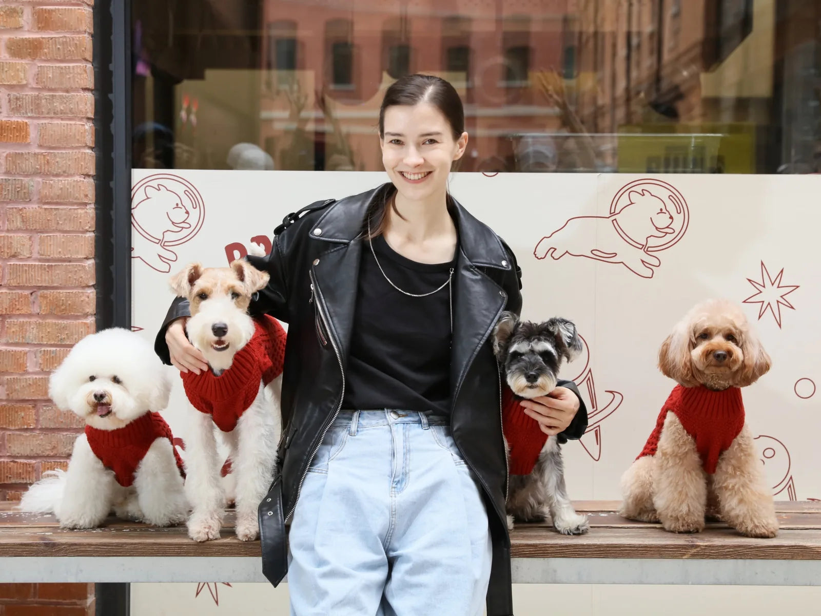 Four dogs in cozy red sweater vests are being held by a smiling woman, showcasing stylish pet apparel against a fun backdrop.