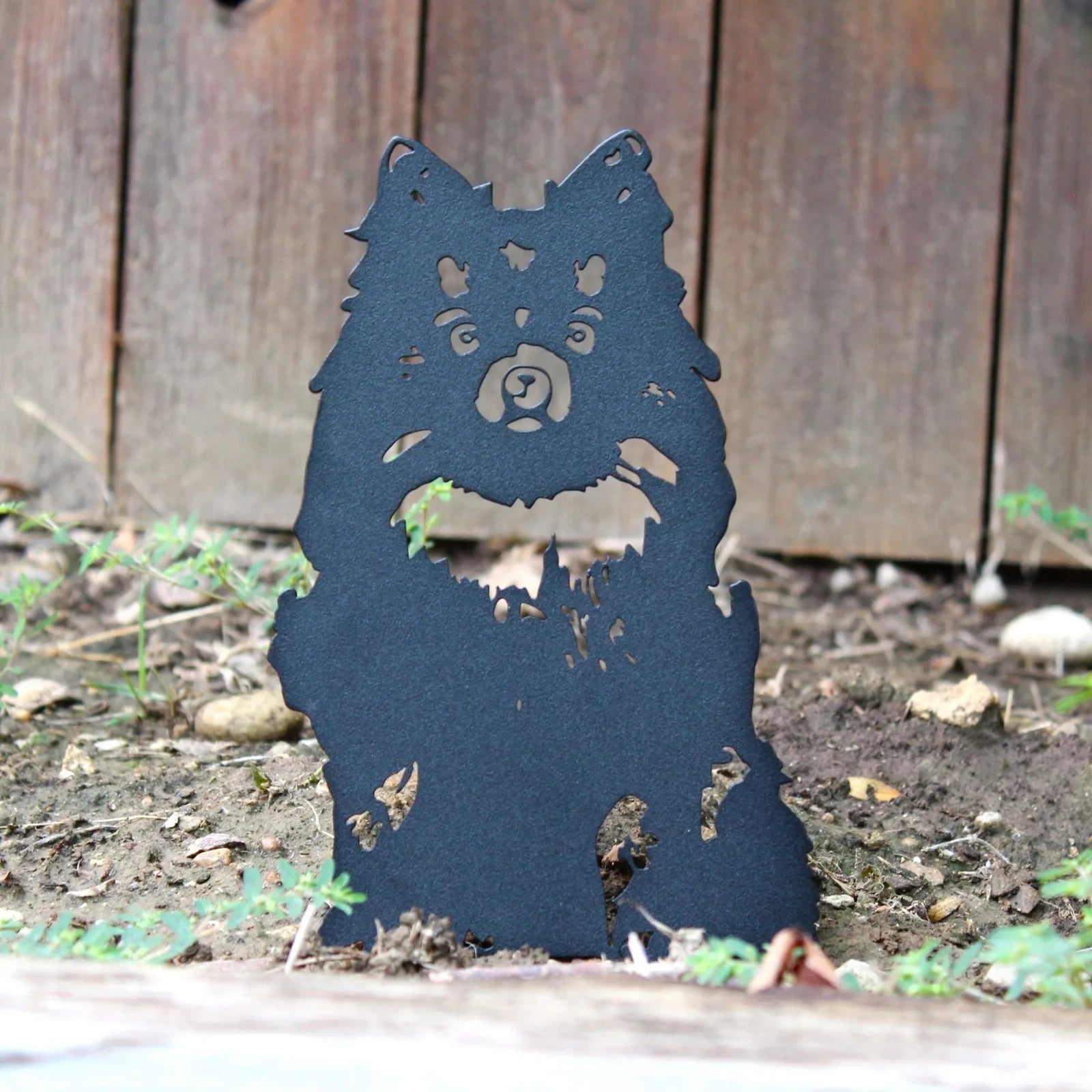 Black metal silhouette of a Pomeranian sitting, nestled in soil with greenery, set against a wooden backdrop.