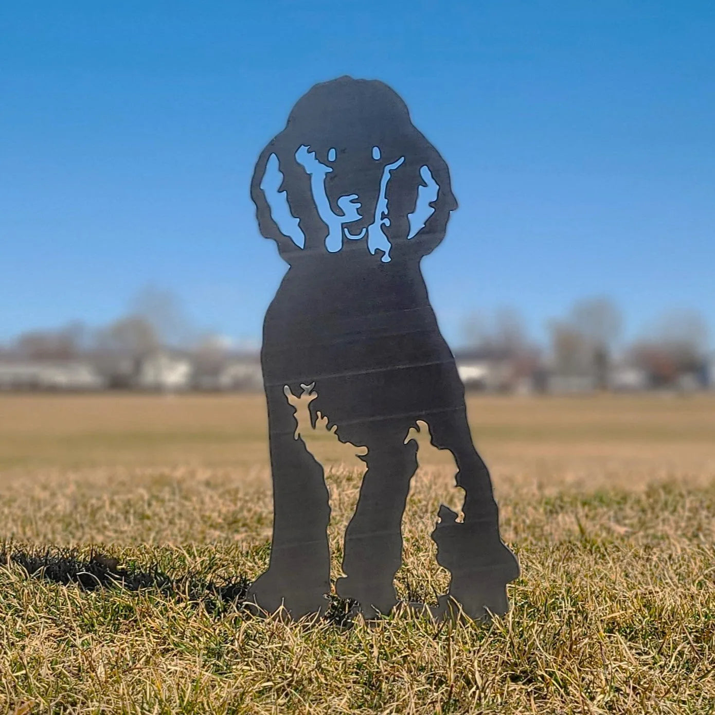 Black metal silhouette of a poodle dog standing on grass against a clear blue sky, ideal for outdoor decor.