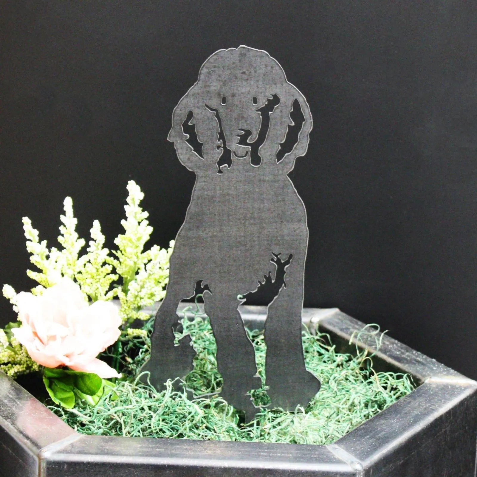 Black metal silhouette of a poodle dog, featured in a decorative plant pot with greenery and a pink flower.