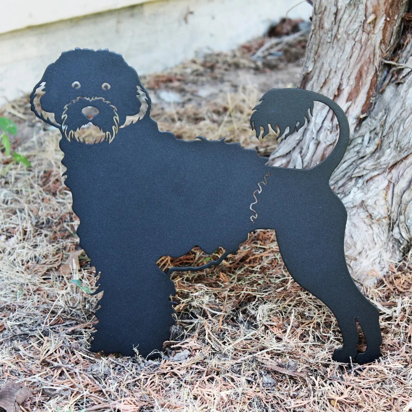 Black metal silhouette of a Portuguese Water Dog, featuring playful details and standing among dried grass and a tree trunk.