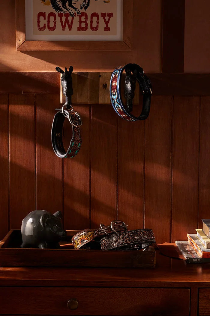 Rustic leather dog collars of various designs hanging on hooks with a pig figurine on a wooden table in a warm-lit room.
