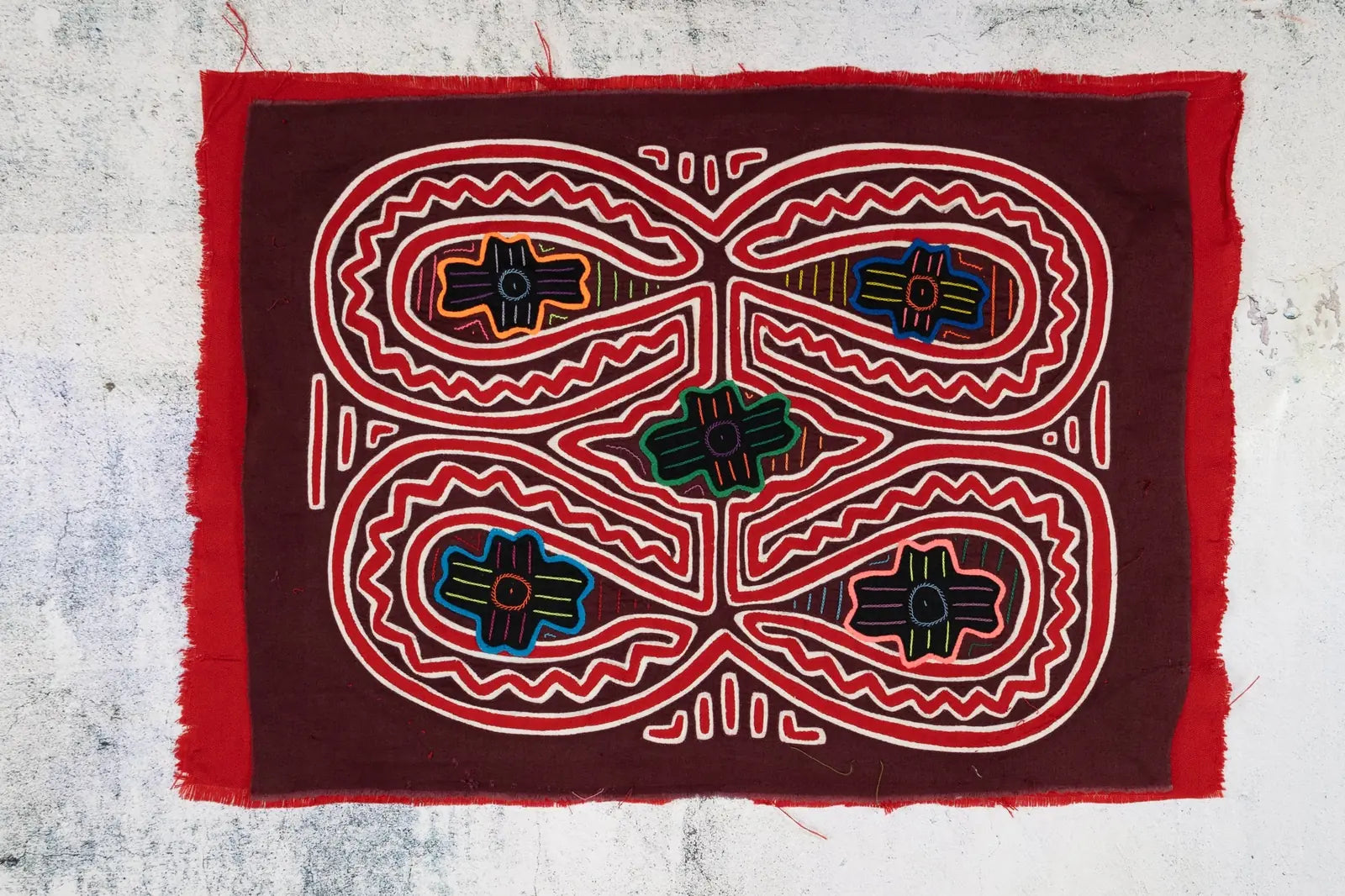 Hand-stitched Mola tapestry featuring intricate geometric patterns on maroon fabric with colorful accents.