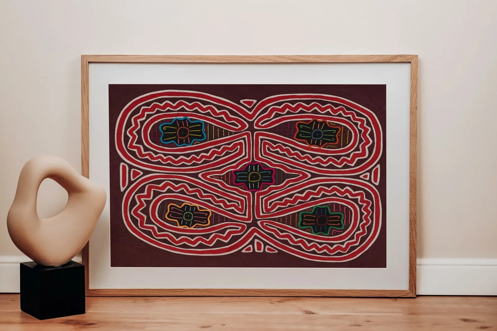 Colorful hand-stitched textile featuring geometric patterns on a deep maroon background, framed and displayed beside a mod...