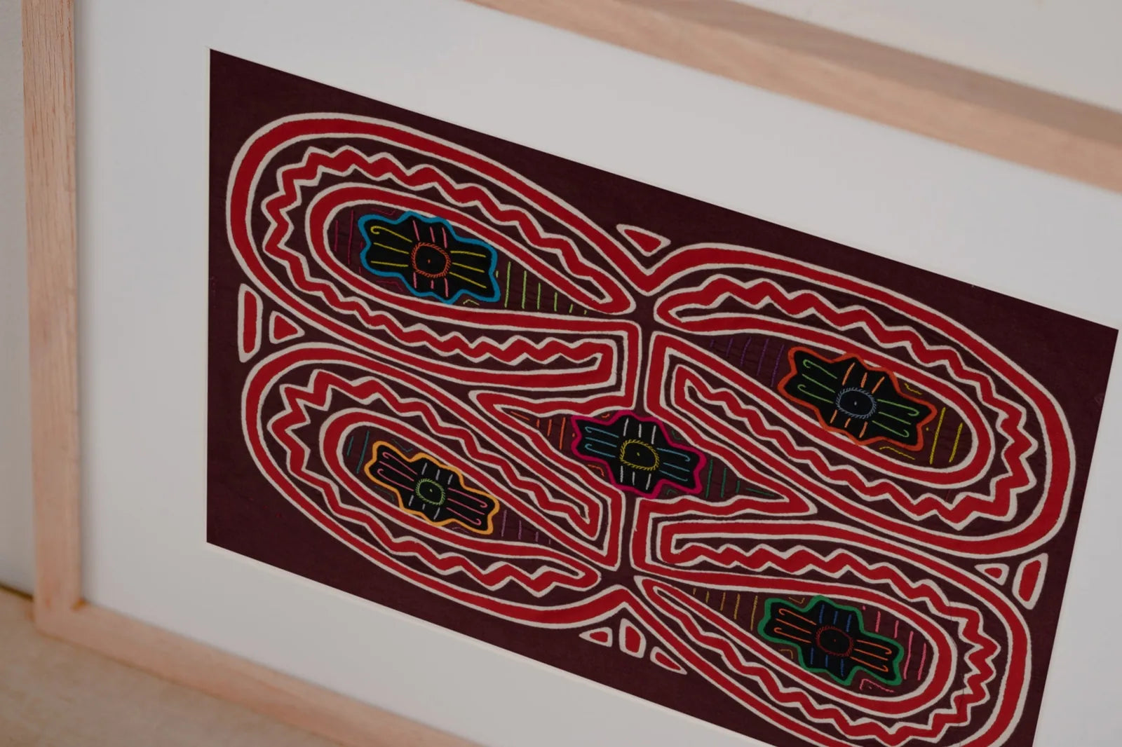 Colorful hand-stitched Mola textile art featuring geometric patterns on a deep maroon background, framed elegantly.
