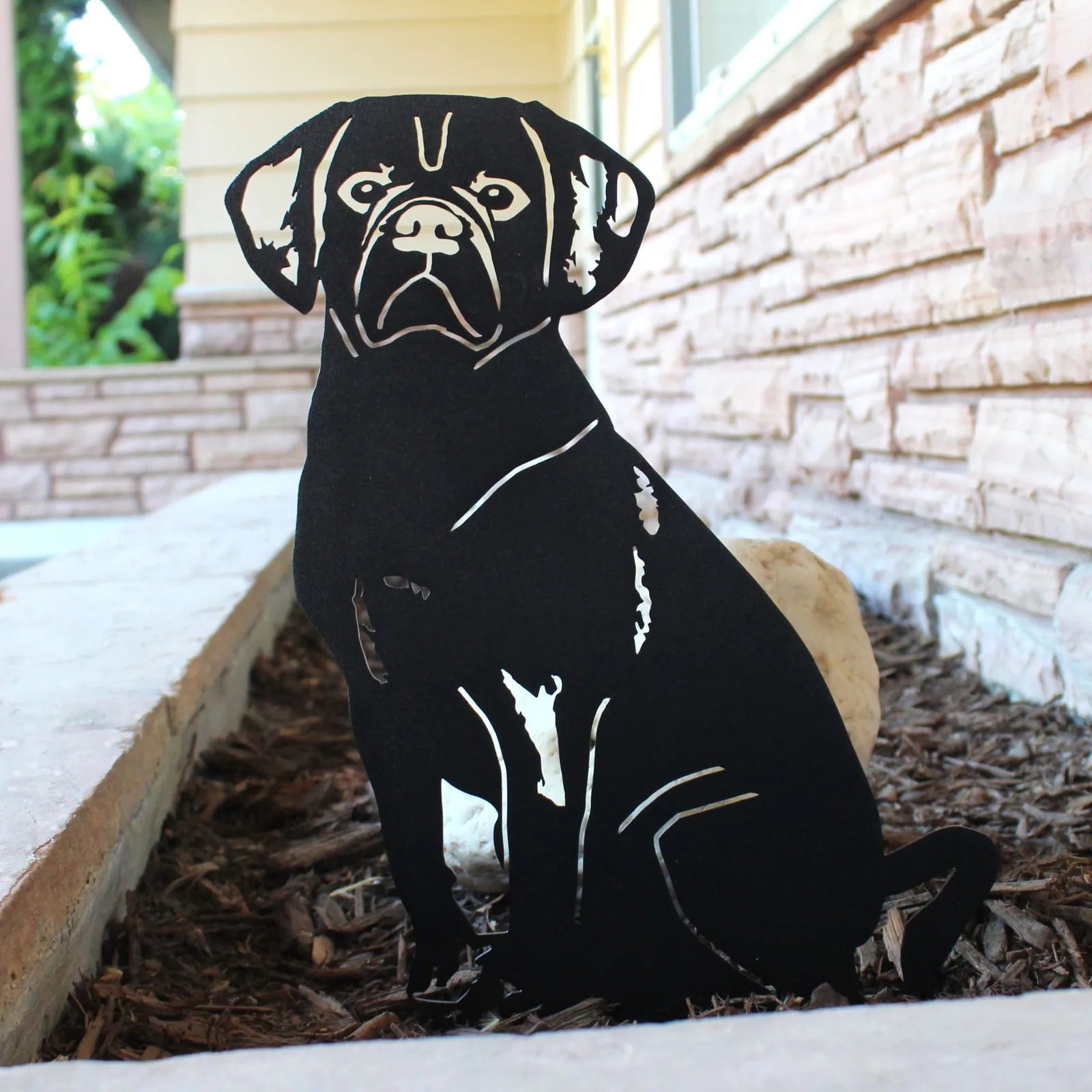 Black Puggle metal silhouette with intricate cutouts, set in mulch beside a stone wall, showcasing detailed craftsmanship.