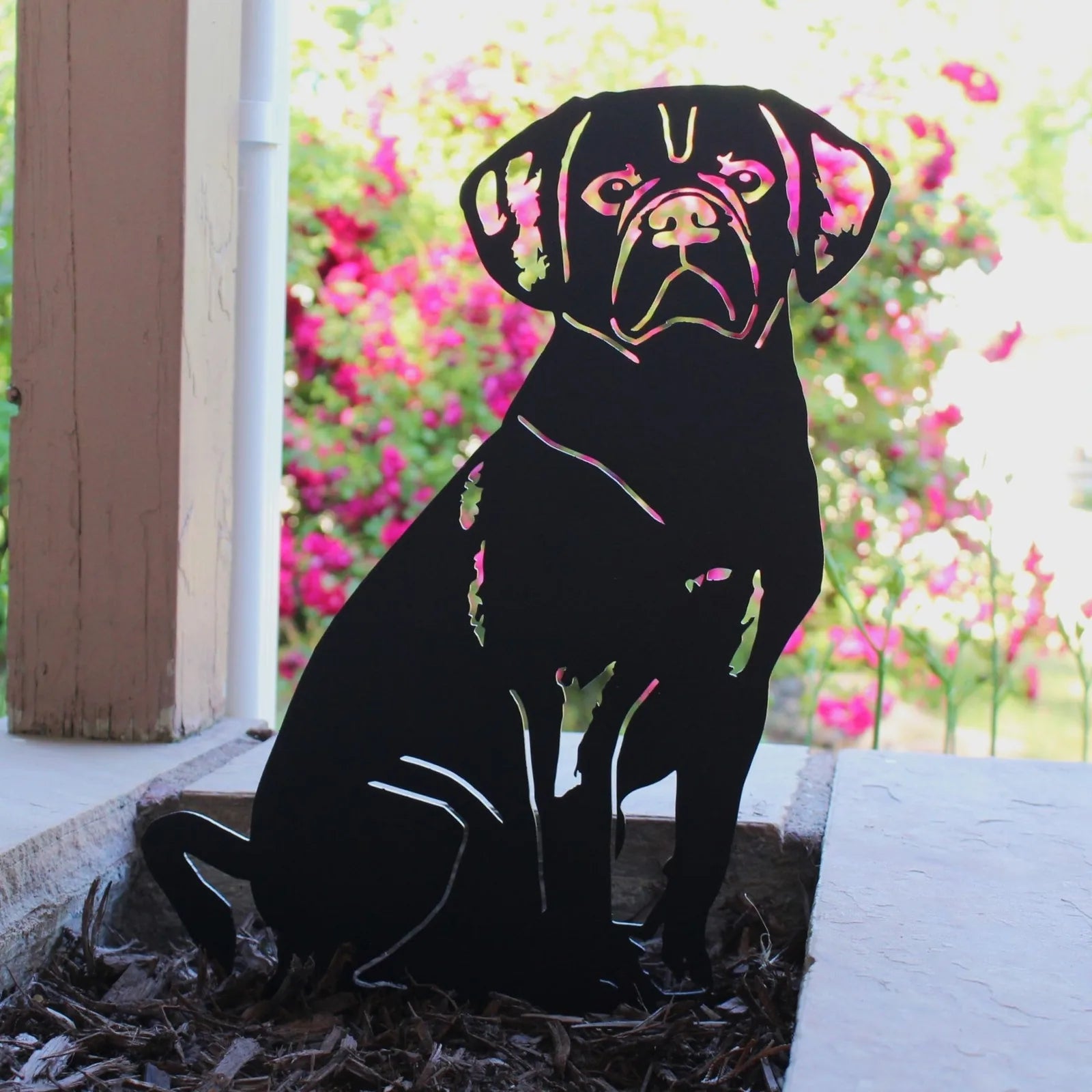 Black metal silhouette of a sitting Puggle with a glossy finish, surrounded by vibrant garden flowers.