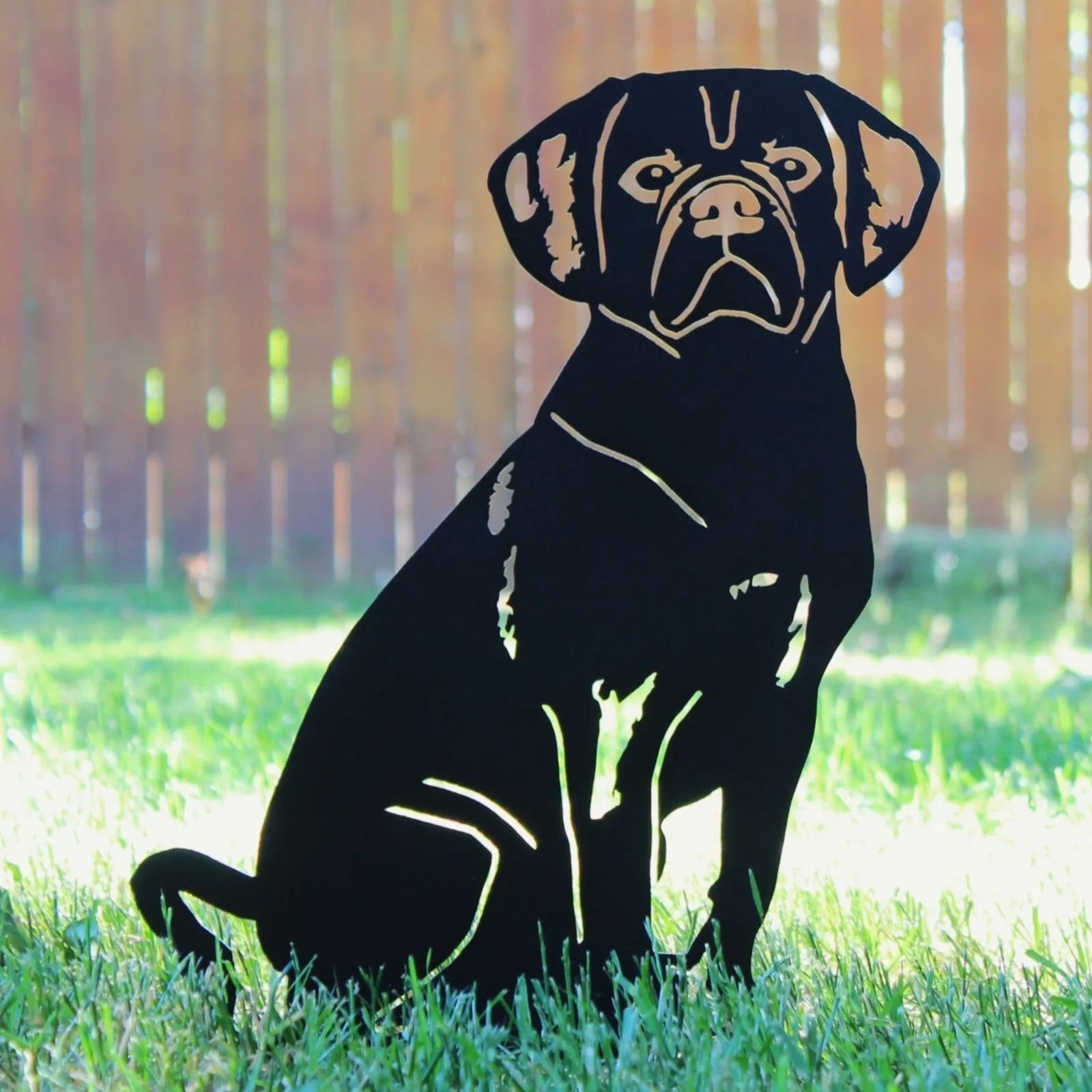 Handmade black metal silhouette of a seated Puggle dog on green grass, set against a wooden fence background.