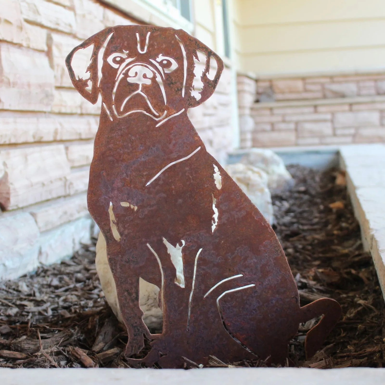 Rusty Corten steel Puggle silhouette, featuring intricate detailing, placed in garden mulch against a stone backdrop.