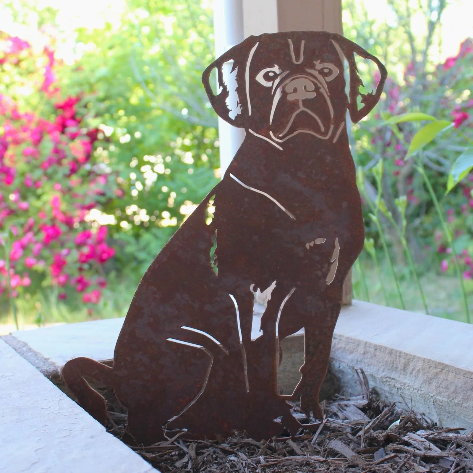 Handmade Corten steel Puggle silhouette with rust patina, placed in a garden setting surrounded by greenery and flowers.