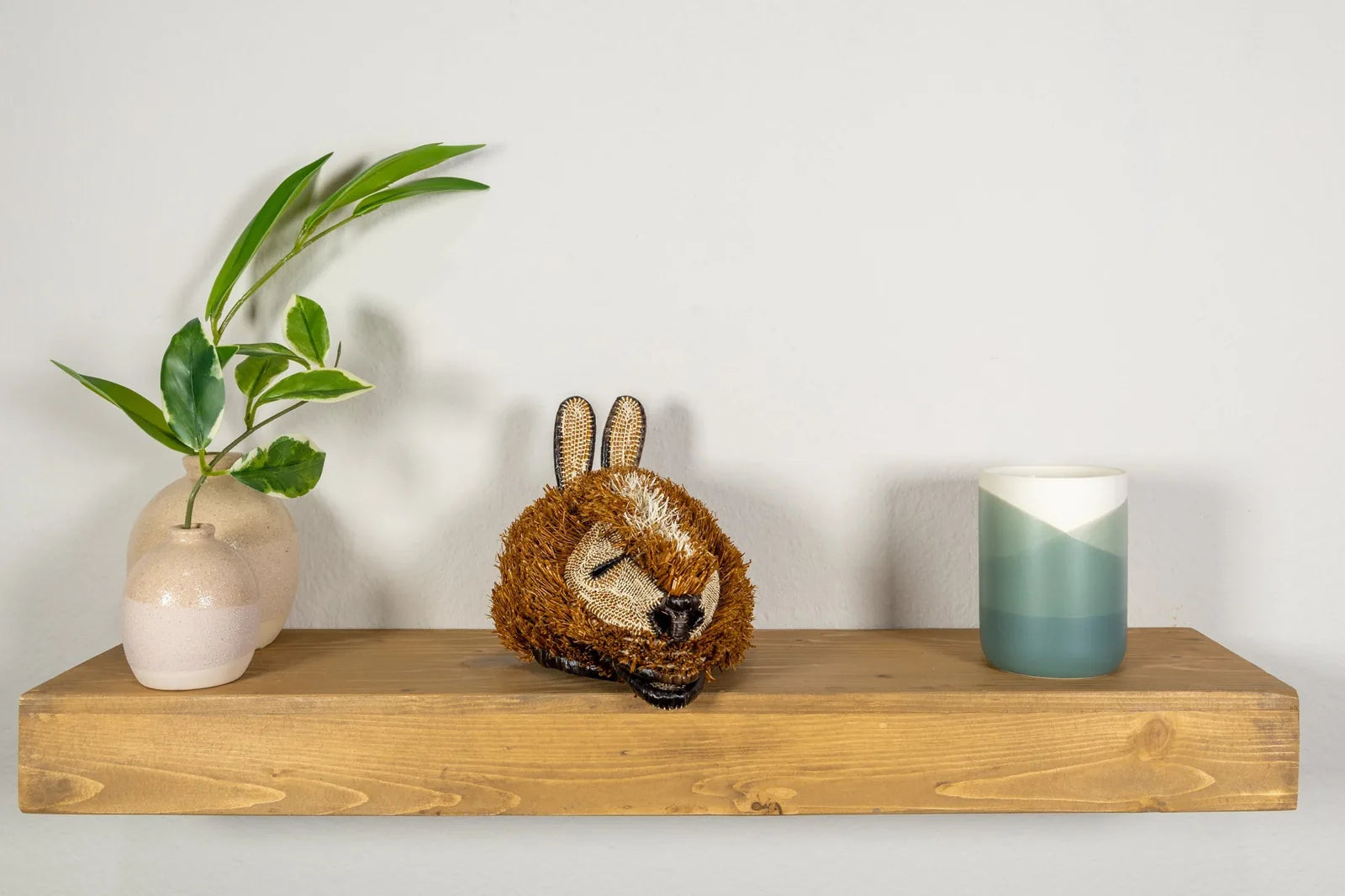 Handwoven Puppy Dog Mask in earthy browns, displayed on a wooden shelf with vases and a candle.