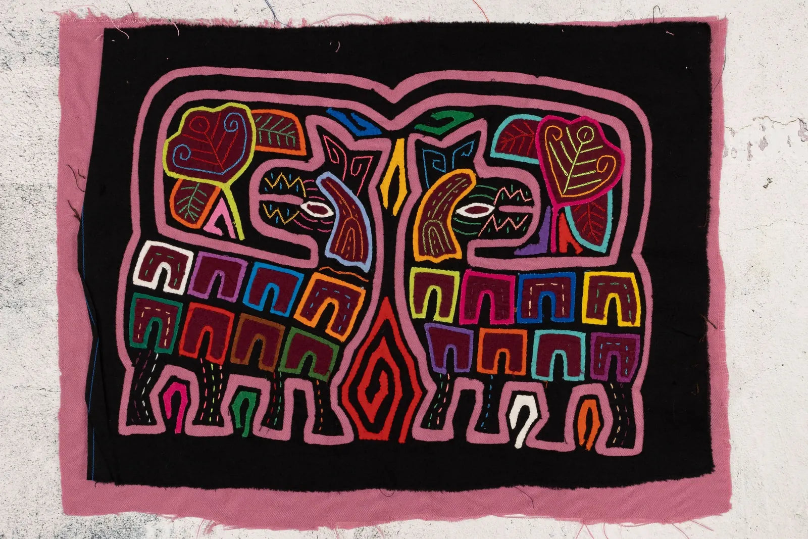Colorful, hand-stitched Mola featuring two geometric dogs amidst vibrant leaves on a black background, framed in pink fabric.