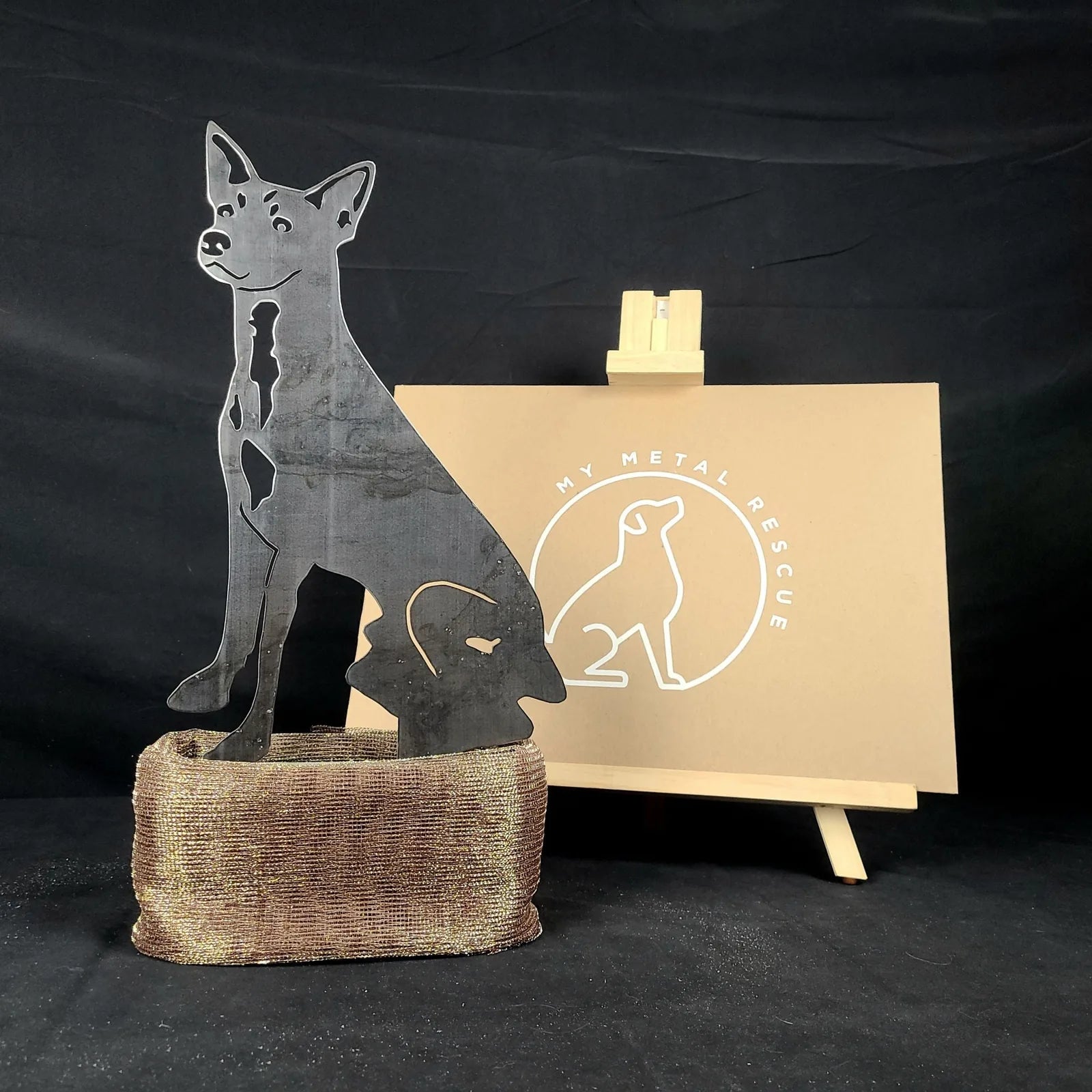Rat Terrier metal silhouette, crafted from black Corten steel, displayed on a rustic brown base with branding backdrop.
