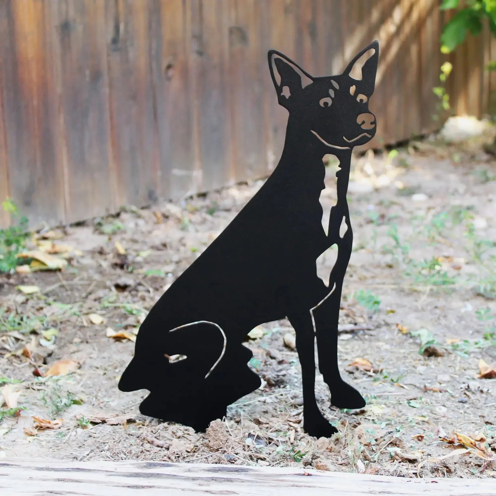 Black metal silhouette of a Rat Terrier sitting, showcasing intricate details, against a natural outdoor setting.